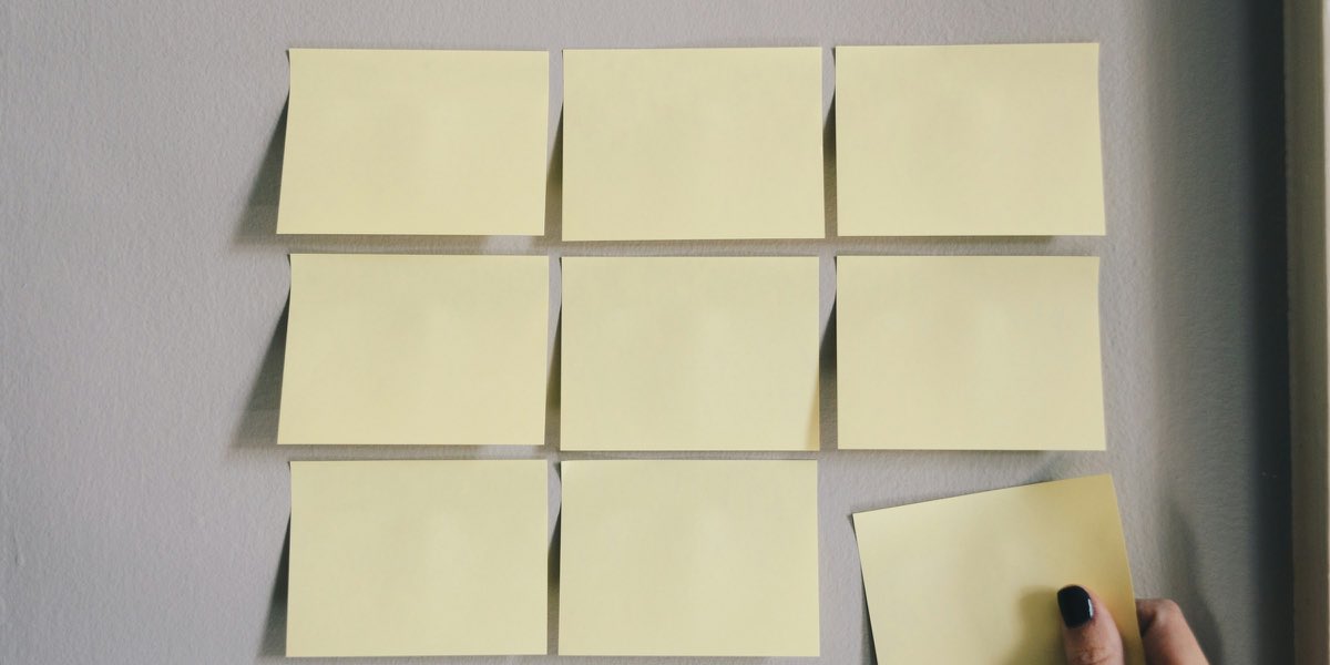 A grid of blank yellow sticky notes on a wall, one being placed by hand